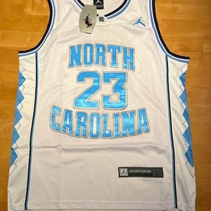 May include: White basketball jersey with blue and silver accents. The jersey has "NORTH CAROLINA" and the number "23" on the front. The Jordan logo is on the right shoulder and the Jordan brand tag is on the bottom right.