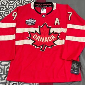 May include: A red hockey jersey with white stripes on the sleeves and shoulders. The jersey features the word "CANADA" within a maple leaf emblem on the chest. The numbers "9" and "7" are on the sleeves. The jersey has the text "NATIONS FACE-OFF 2022".