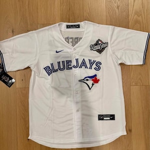 Może przedstawiać: Biała koszulka baseballowa z niebieskimi i czerwonymi akcentami. Zawiera napis "BLUEJAYS" w niebieskim kolorze na piersi, logo ptaka i liścia klonu oraz naszywkę "World Series". Widoczne jest logo Nike.