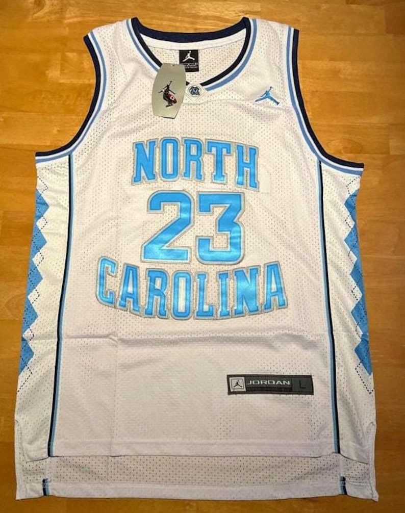 K&ouml;nnte beinhalten: Wei&szlig;es Basketballtrikot mit den Worten "NORTH CAROLINA" und der Nummer "23" in Hellblau. Das Trikot hat einen blauen Besatz und ein Rautenmuster an den Seiten. Ein "JORDAN"-Etikett ist sichtbar.