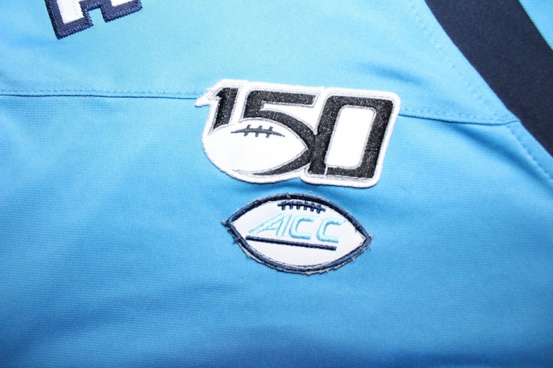 May include: A light blue jersey with a black and white patch that says "150" and a white patch with a football and the text "ACC".