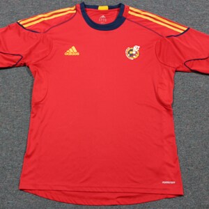 May include: Red soccer jersey with navy blue trim and yellow and gold stripes on the shoulders. The Adidas logo is on the left chest and the Spanish national team crest is on the right chest. The text "PERFORMANCE" is on the bottom right of the jersey.
