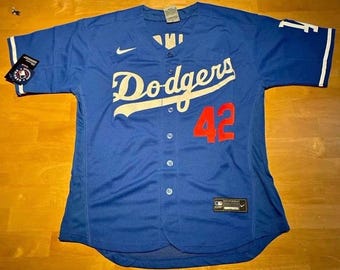 May include: A blue Dodgers baseball jersey with white lettering and the number 42 in red. The jersey has a Nike logo and a tag on the sleeve. The jersey is button-down and has white trim on the sleeves.