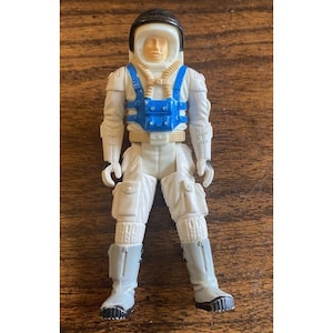 May include: A vintage astronaut action figure in a white spacesuit with a blue harness. The figure has a black helmet and grey boots. The toy is standing on a wooden surface.
