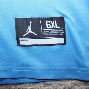 May include: A light blue t-shirt with a black tag that says "6XL" and "Quality Performance Product Inspired by the Greatest Player Ever".