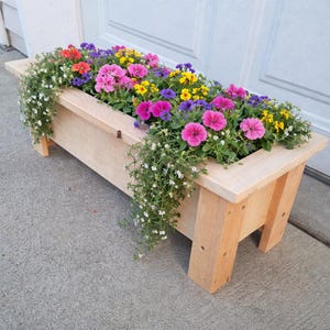 May include: A rectangular wooden planter box filled with vibrant pink, yellow, purple, and red flowers. Trailing white flowers cascade over the sides. The planter has a natural wood finish and is supported by angled legs.