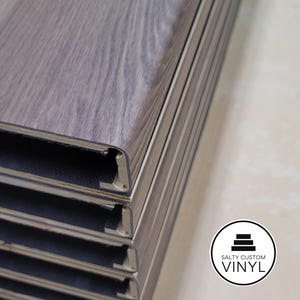 May include: Close-up of stacked gray vinyl flooring planks made into custom stair treads with a wood grain pattern. The planks are interlocking and have a beveled edge. The Salty Custom Vinyl logo is in the bottom right corner.