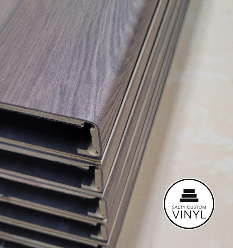 May include: Close-up of stacked gray vinyl flooring planks made into custom stair treads with a wood grain pattern. The planks are interlocking and have a beveled edge. The Salty Custom Vinyl logo is in the bottom right corner.