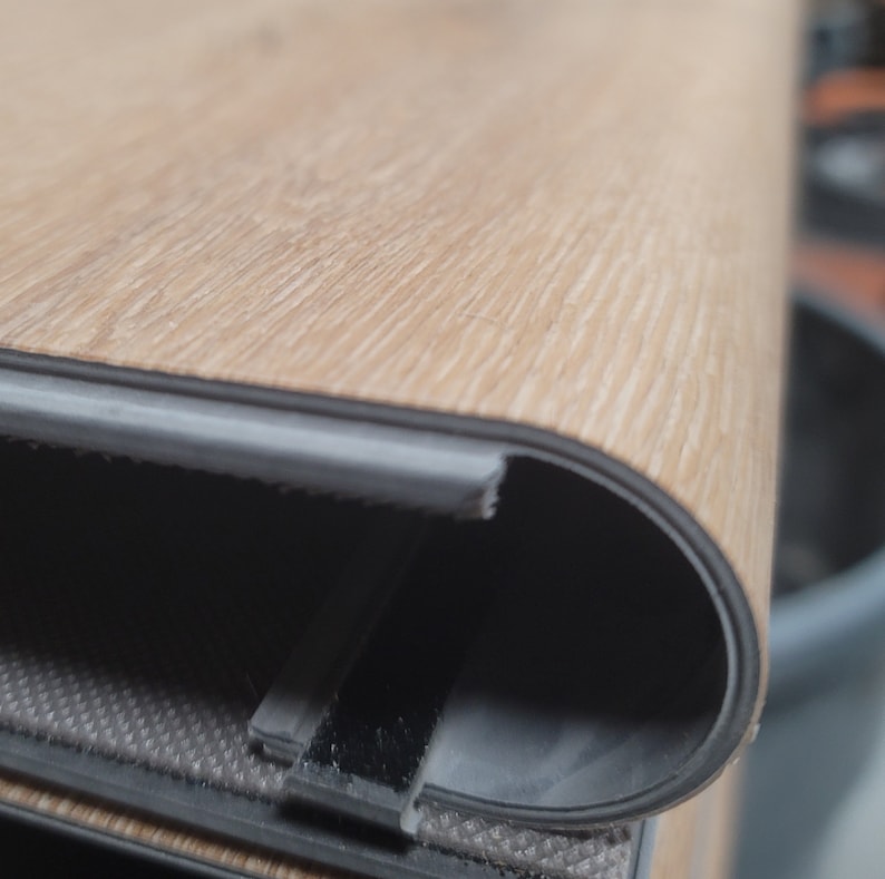 May include: Close-up of layered flooring material. The top layer has a light brown wood grain pattern. The edges are made up of a locking system mechanism, revealing the layers of the plank column. The flooring appears to be new and unused.