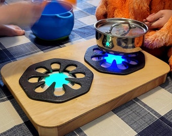 Puede incluir: Una estufa de juguete de madera con dos anillos de quemador de metal negro. Cada anillo de quemador tiene una luz azul que simula una llama. Una olla de plata con tapa se encuentra sobre uno de los anillos de quemador.