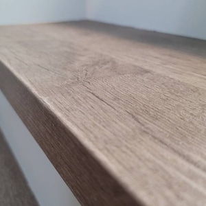 May include: Close-up of a luxury vinyl custom stair tread with a light brown, wood textured surface. The stair tread is mounted on a staircase tread step and against a light blue wall. The wood grain is visible.