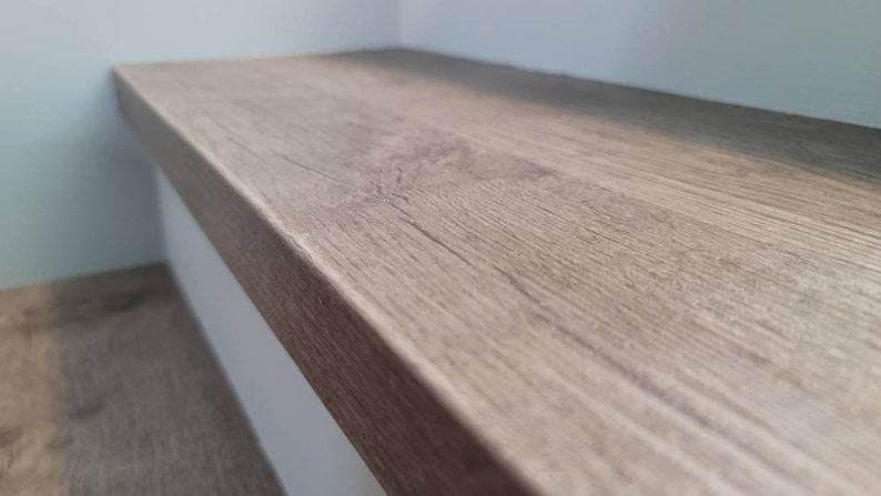 May include: Close-up of a luxury vinyl custom stair tread with a light brown, wood textured surface. The stair tread is mounted on a staircase tread step and against a light blue wall. The wood grain is visible.