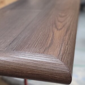 May include: Close-up of a dark brown wooden looking stair step with a beveled edge. The wood grain is visible, and the step appears to be well-crafted. The corner of the step is rounded with multiple finished sides.