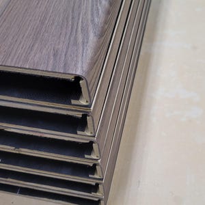 May include: A stack of brownish wood-look flooring planks shaped into stair tread covers. The planks have a textured surface with a wood grain pattern. The edges of the planks show a plank core made of wpc. The vinyl flooring is a neutral color.