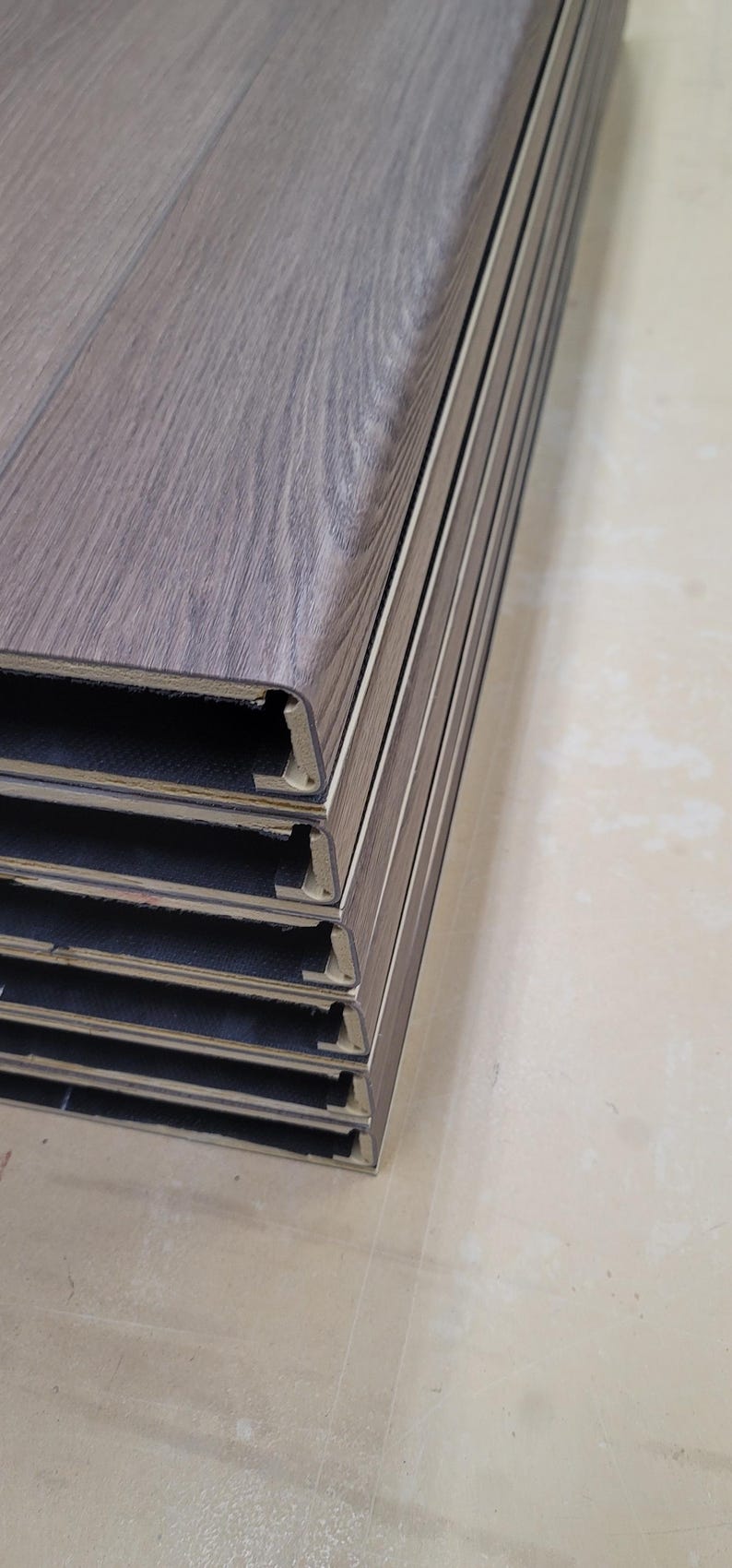 May include: A stack of brownish wood-look flooring planks shaped into stair tread covers. The planks have a textured surface with a wood grain pattern. The edges of the planks show a plank core made of wpc. The vinyl flooring is a neutral color.