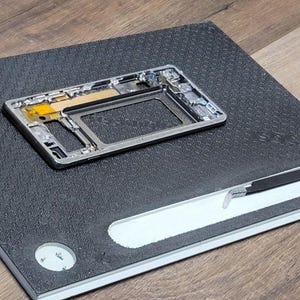 May include: A disassembled smartphone frame with internal components rests on a textured black surface. A white and black tool is placed on a white section of the surface. The background is a wooden surface.