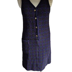 May include: A sleeveless, knee-length dress with a V-neck and a geometric pattern in shades of purple, black, and grey. The dress features gold-coloured buttons down the front and a small pocket on the left side. The dress is displayed on a mannequin.