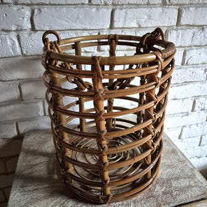 Può includere: Un cesto cilindrico in rattan a trama aperta. Il cesto è di colore marrone caldo e ha una struttura robusta e intrecciata. Presenta una parte superiore arrotondata con manici ed è adatto per la conservazione o per scopi decorativi.