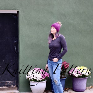May include: A woman wearing a purple knitted hat with a white pom pom, a grey long-sleeved shirt, and blue jeans. She is standing in front of a green wall with a black door.