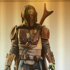 Puede incluir: Ilustraci&oacute;n de un guerrero mandaloriano con armadura detallada. El casco es verde oscuro con una visera en forma de T. La armadura es una mezcla de marr&oacute;n, bronce y verde, con una capa y una pistola bl&aacute;ster. Una lanza est&aacute; unida a la espalda.