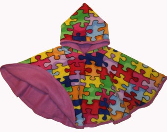 May include: A children's hooded poncho with a vibrant jigsaw puzzle design in various colours. The interior and trim are a solid pink. The poncho is made from a soft, fleece-like fabric, perfect for keeping warm.