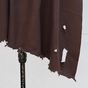 Puede incluir: Una tela marr&oacute;n oscuro con un borde desgastado y rasgado y varios agujeros. La tela est&aacute; drapeada sobre un soporte negro, creando una apariencia fluida y texturizada. El material parece ser de peso pesado.