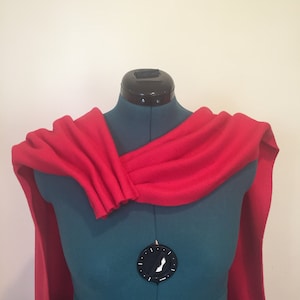 Puede incluir: Una capa roja vibrante drapeada sobre un maniqu&iacute; turquesa. La capa est&aacute; hecha de una tela suave y fluida, con un detalle fruncido en el hombro. La longitud de la capa se extiende por el maniqu&iacute;, creando una silueta dram&aacute;tica. El fondo es una pared lisa blanquecina.