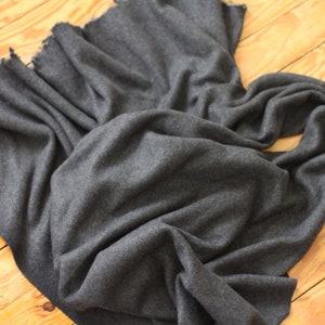 Puede incluir: Una bufanda gris oscuro hecha de tela suave, drapeada sobre una superficie de madera. La bufanda tiene un borde con flecos y est&aacute; doblada de manera que muestra su textura y ca&iacute;da. El aspecto general es simple y elegante.