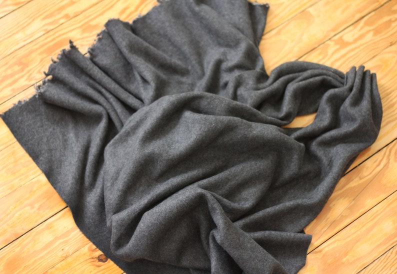 Puede incluir: Una bufanda gris oscuro hecha de tela suave, drapeada sobre una superficie de madera. La bufanda tiene un borde con flecos y est&aacute; doblada de manera que muestra su textura y ca&iacute;da. El aspecto general es simple y elegante.