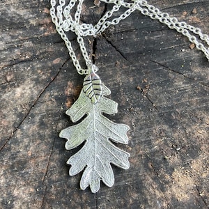 May include: Silver-toned necklace featuring a detailed oak leaf pendant. The pendant is suspended from a delicate chain, showcasing the intricate veining of the leaf. The necklace is displayed on a rustic wooden surface.