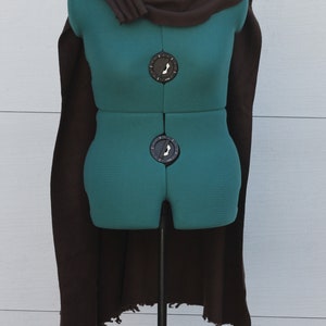 Puede incluir: Una capa marr&oacute;n drapeada sobre un maniqu&iacute; verde. La capa tiene un cuello alto y bordes deshilachados. El maniqu&iacute; est&aacute; sobre un soporte met&aacute;lico. La capa est&aacute; hecha de una tela suave. El maniqu&iacute; se utiliza para exhibir ropa.