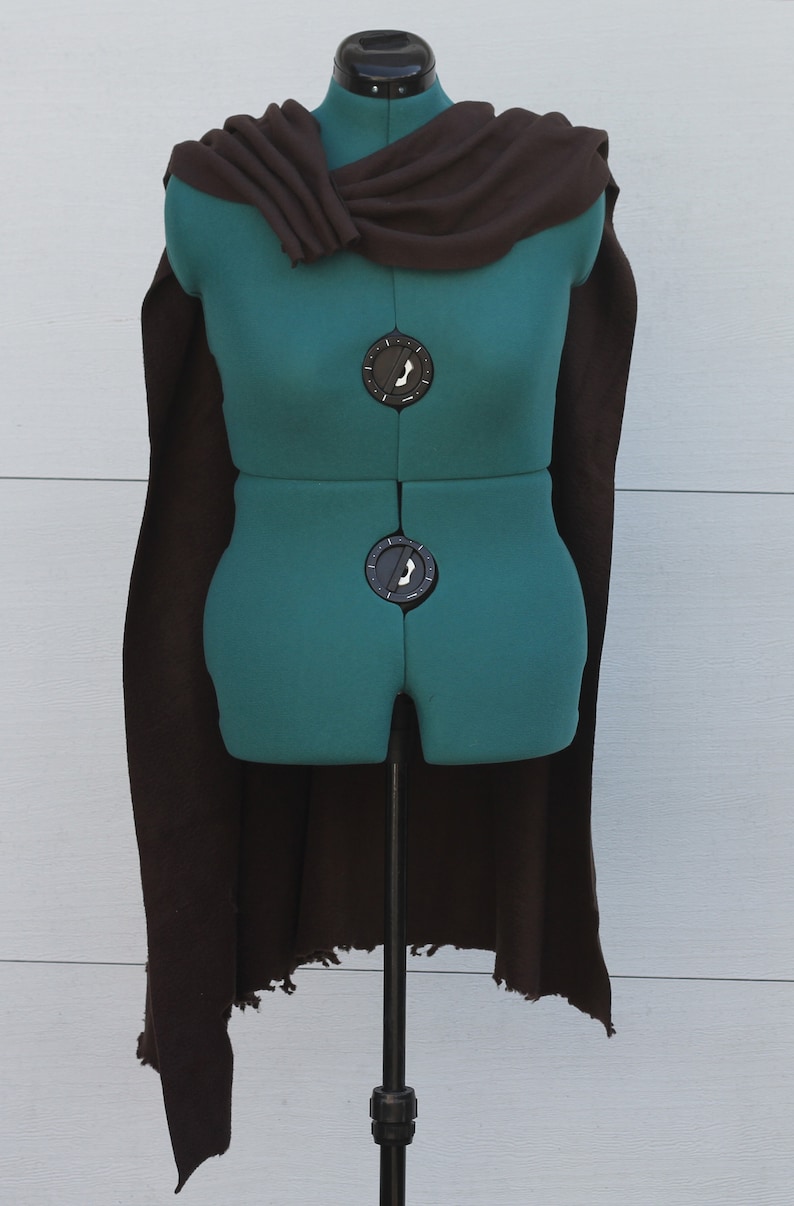 Puede incluir: Una capa marr&oacute;n drapeada sobre un maniqu&iacute; verde. La capa tiene un cuello alto y bordes deshilachados. El maniqu&iacute; est&aacute; sobre un soporte met&aacute;lico. La capa est&aacute; hecha de una tela suave. El maniqu&iacute; se utiliza para exhibir ropa.