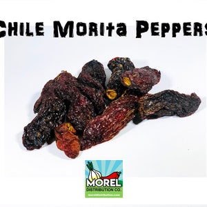 Può includere: Peperoncini morita secchi, un tipo di peperoncino secco, sono mostrati in un mucchio su una superficie bianca. I peperoncini sono di colore rosso scuro e rugosi. L'immagine include anche un logo per Morel Distribution Co.
