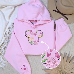 May include: Pink hoodie with a floral Mickey Mouse design embroidered on the front. The design includes pink, yellow, and green flowers, as well as small pink hearts.
