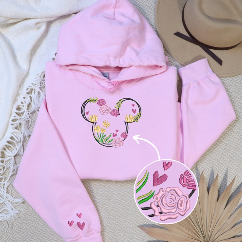 May include: Pink hoodie with a floral Mickey Mouse design embroidered on the front. The design includes pink, yellow, and green flowers, as well as small pink hearts.