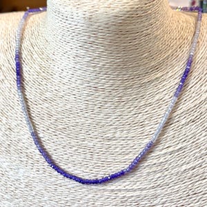 May include: A delicate necklace featuring small, faceted beads in shades of purple and white. The beads transition from a deep purple to a lighter, almost clear color, creating an ombre effect. The necklace is displayed on a neutral-toned bust.