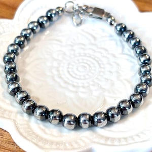 May include: A beaded bracelet featuring polished, dark gray spherical beads. The bracelet is strung on a flexible cord and secured with a silver-colored clasp. The beads are approximately 8mm in diameter. The bracelet is displayed on a white, decorative plate.