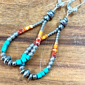 May include: A pair of handcrafted earrings with a teardrop design. The earrings feature turquoise, orange, and red beads, interspersed with silver-colored metal beads. The earrings have a silver-colored hook closure.
