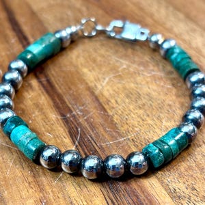 May include: A beaded bracelet featuring alternating polished silver beads and vibrant turquoise-green stone beads. The bracelet is strung on a silver chain and secured with a silver clasp. The bracelet is displayed on a wooden surface.