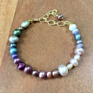 May include: A rainbow gradient pearl bracelet with a gold clasp. The pearls are iridescent and range in color from green to purple to pink to white.