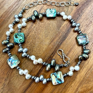 May include: A necklace featuring alternating white and dark gray freshwater pearls, interspersed with square-shaped iridescent abalone shell beads and small metallic spacer beads. The piece includes a silver-toned hook clasp and an adjustable chain.