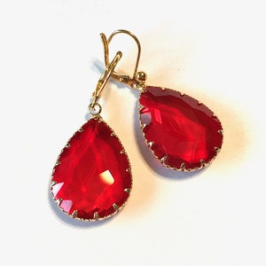 May include: A pair of gold earrings with red teardrop-shaped crystals. The crystals have a faceted design and are set in a gold frame.