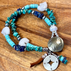 May include: A turquoise and white beaded bracelet with a silver compass charm, a buffalo nickel charm, and a brown leather tassel. The bracelet is made with turquoise, white, and silver beads.