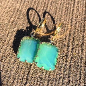 May include: A pair of gold hoop earrings with light green rectangular gemstones. The earrings have a delicate, ornate design.