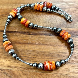 May include: A beaded necklace featuring alternating sections of polished metallic silver-toned spheres and vibrant orange, disc-shaped beads with marbled white patterns. The strand is secured with a metallic silver-toned lobster claw clasp.