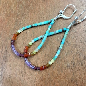 May include: A pair of dangle earrings with a gradient of gemstone beads. The beads are in shades of purple, red, orange, yellow, and blue. The earrings are made with silver hooks.