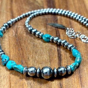 May include: Silver-toned necklace with turquoise and silver beads. The necklace features a mix of round silver beads and irregular turquoise stones, creating a striking contrast. The necklace is displayed on a wooden surface.