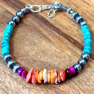 May include: A beaded bracelet featuring turquoise, silver, and colorful gemstone beads. The bracelet includes a mix of round silver beads, vibrant turquoise, and a section of orange, pink, and purple stones. It has a silver clasp.