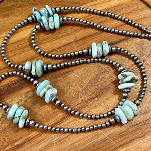 May include: A silver beaded necklace with turquoise-colored stone accents. The necklace features multiple strands of small, round silver beads interspersed with larger, irregular turquoise stones. The necklace is displayed on a wooden surface.