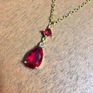 May include: A gold chain necklace with a teardrop-shaped red gemstone pendant and a smaller square red gemstone above it.